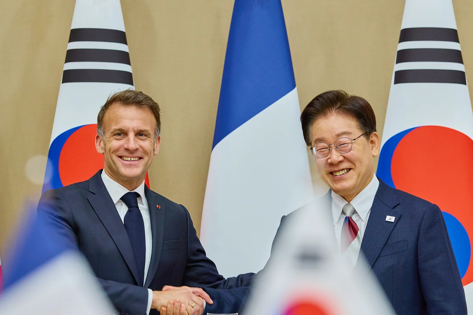 Emmanuel Macron et Lee Jae-myung se serrent la main lors du sommet France-Corée du Sud, le 3 avril 2026 à Séoul, devant les drapeaux français et sud-coréen.