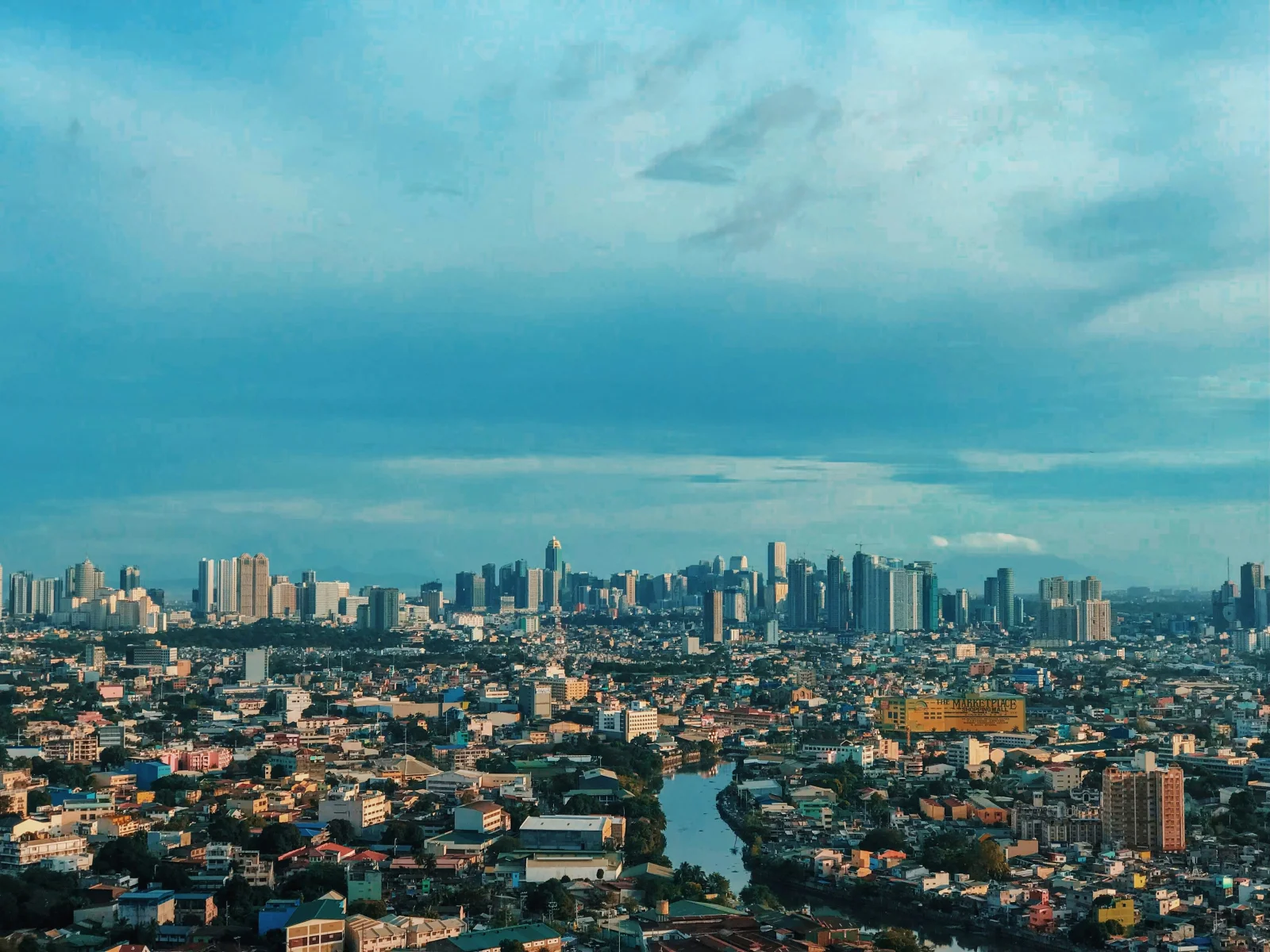 Vue aérienne panoramique de Manille, Philippines, sous un ciel bleu nuageux, avec une rivière sinuant entre des quartiers résidentiels denses et la skyline du Makati CBD en arrière-plan.